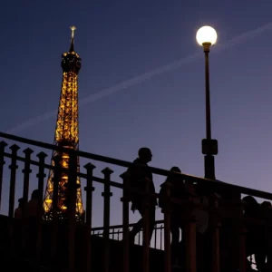 People walk on a bridge next to the Eiffel Tower in Paris, Wednesday Feb. 9, 2022. Lights on the Eiffel Tower will soon be turned off an hour earlier at night as part of an energy savings plan in the French capital, its mayor announced. Paris mayor said the iconic tower that is illuminated until 1:00am is only one of the city's monuments and municipal buildings that will be plunged into darkness earlier in the evening as the French capital faces risks of power shortages, rationing and blackouts when energy demand surges this winter.